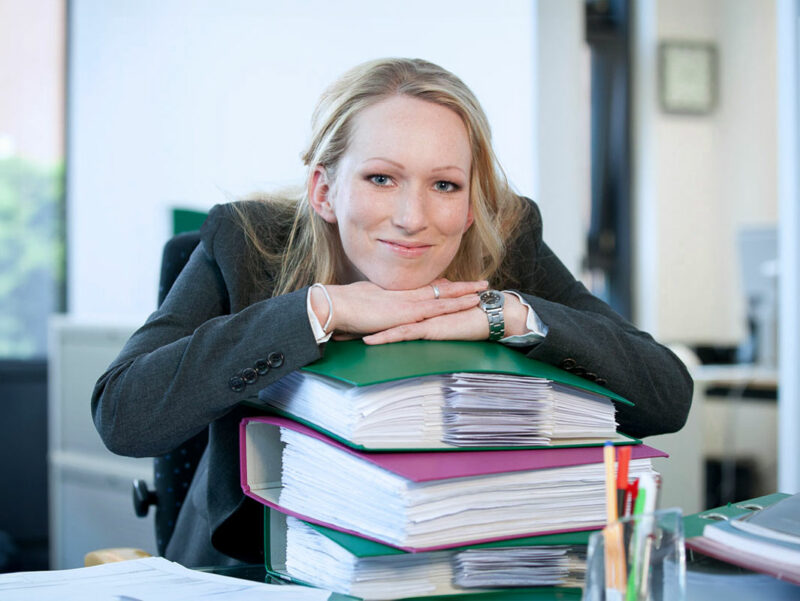 Frau im Büro, stützt sich auf Akten-Ordnern ab. Mitarbeiter Fotos und kreative Portrait Fotografie in Bonn.